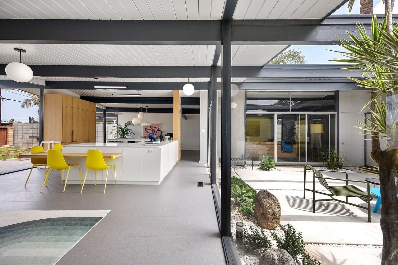 Modern open-plan kitchen and dining area with yellow chairs, white cabinetry, and large windows overlooking a minimalist outdoor courtyard with plants and seating, reflecting why Eichler Homes interior design is a mid-century masterpiece.