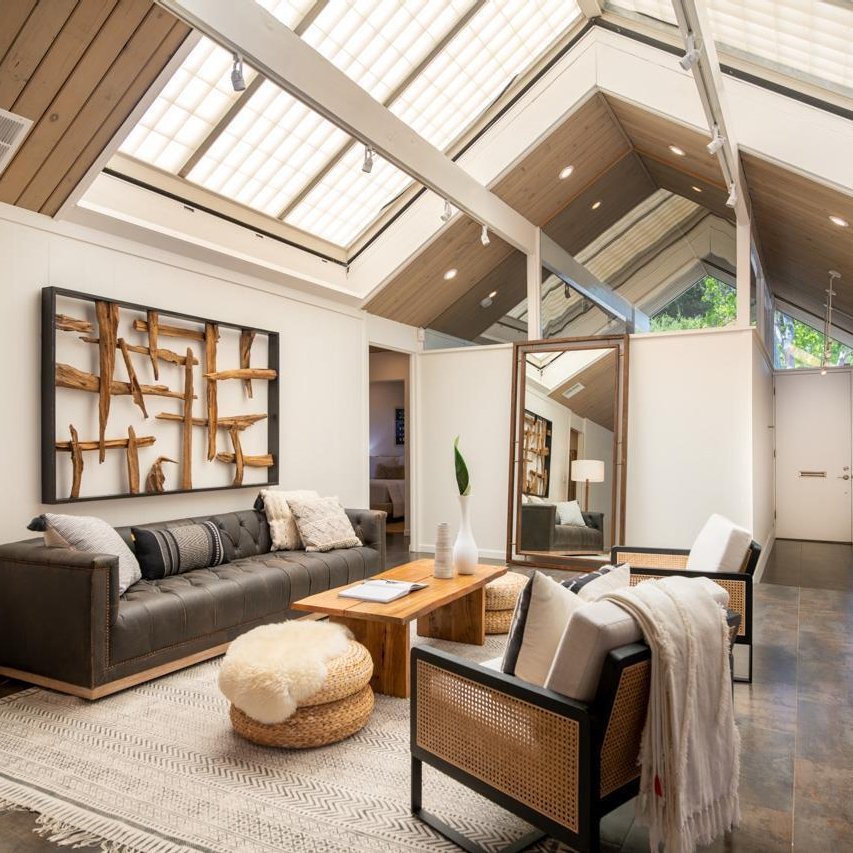 The modern living room features a skylight, black sofas, wooden furniture, a large mirror, and abstract wall art. A rug and poufs add texture, and greenery outside is visible through the windows. It's reminiscent of why Eichler Homes interior design is a mid-century masterpiece.