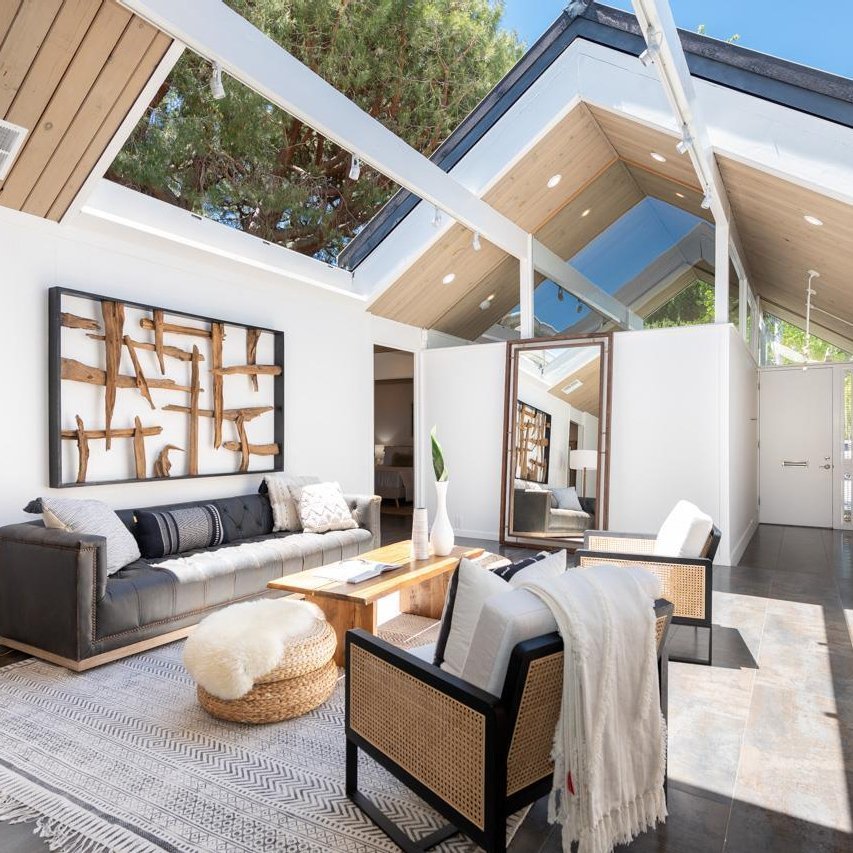 Bright living room with large windows, modern furniture, and wooden decorations. The space echoes why Eichler Homes Interior Design is a mid-century masterpiece, featuring a black sofa, two chairs, a coffee table, and a large mirror against a white wall.