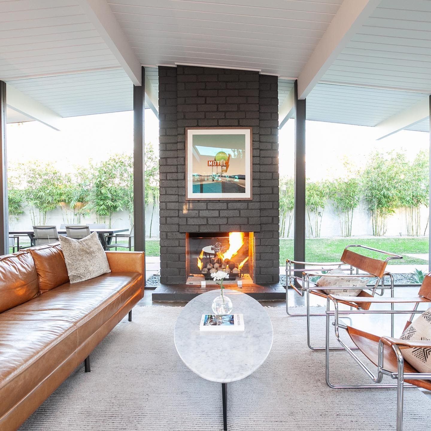 Modern living room with a leather sofa, two sleek armchairs, a round coffee table, and a lit fireplace. Outdoor dining area and lush greenery visible through large windows—truly showcasing why Eichler Homes interior design is a mid-century masterpiece.