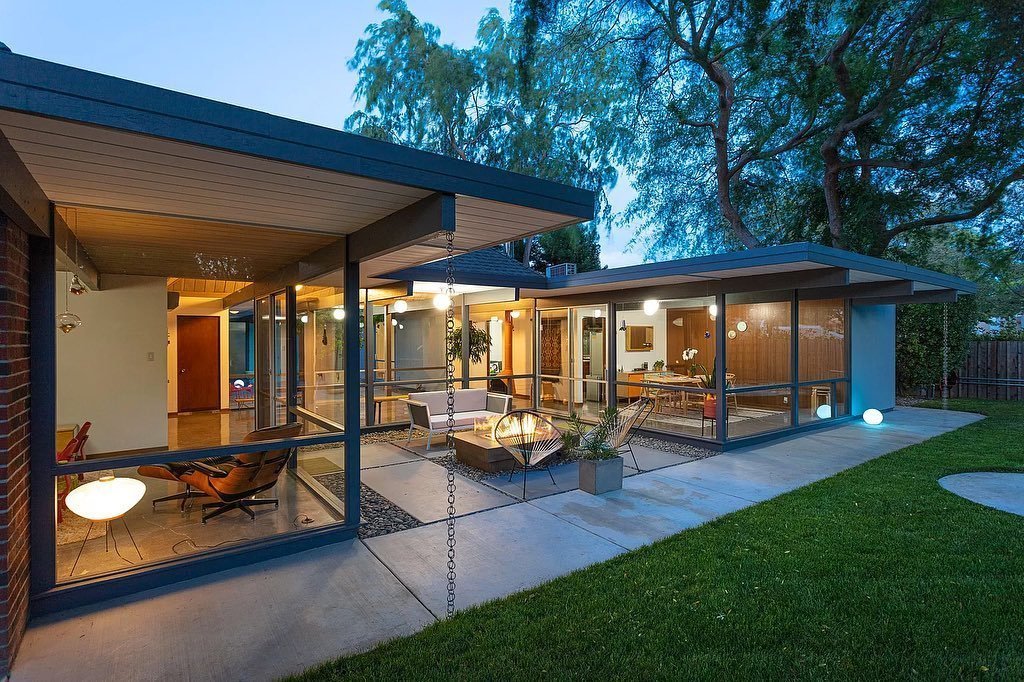 Modern house with extensive glass walls, open living space visible from outside, and outdoor seating area, surrounded by trees and grassy lawn. Evening lighting creates a warm ambiance. Discover why Eichler Homes interior design is a mid-century masterpiece that enhances this inviting atmosphere.