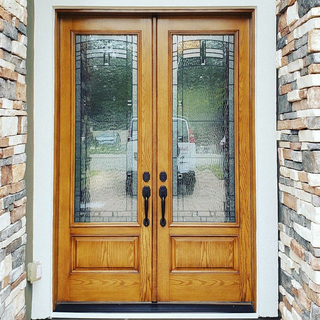 A set of double wooden doors with glass panes framed by a stone wall backdrop. Beyond wood and steel, a vehicle is partially visible through the glass.