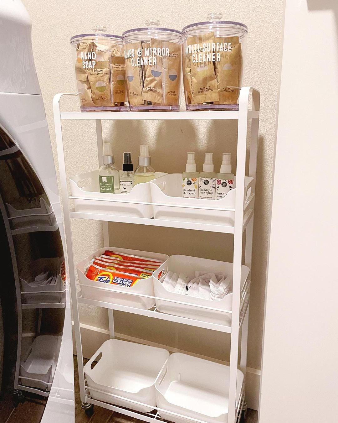 A white, three-tiered storage cart holds hand soap, cleaner, and cleaning supplies in labeled containers, alongside neatly arranged cleaning tools and bottles. Discover how creative storage hacks can transform your laundry area into an organized haven.