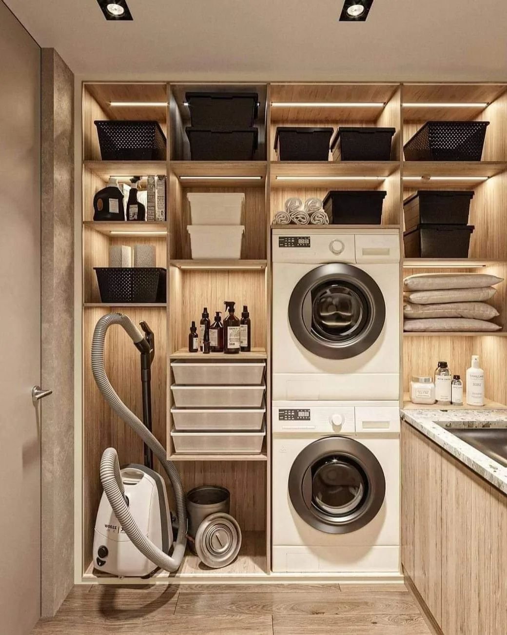 A laundry room features a stacked washer and dryer, shelves with black and white baskets, creative storage hacks for cleaning supplies, a vacuum cleaner, and a counter with neatly folded towels.