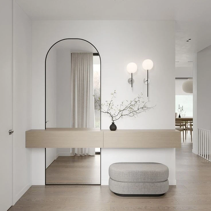 Minimalist entryway with a floating wooden console, a large arched mirror, a small bench, wall-mounted lights, and a vase with branches. Discover how this small entryway makes a big impact with thoughtful design elements inspired by our guide: "10 Design Hacks for Small Foyers.