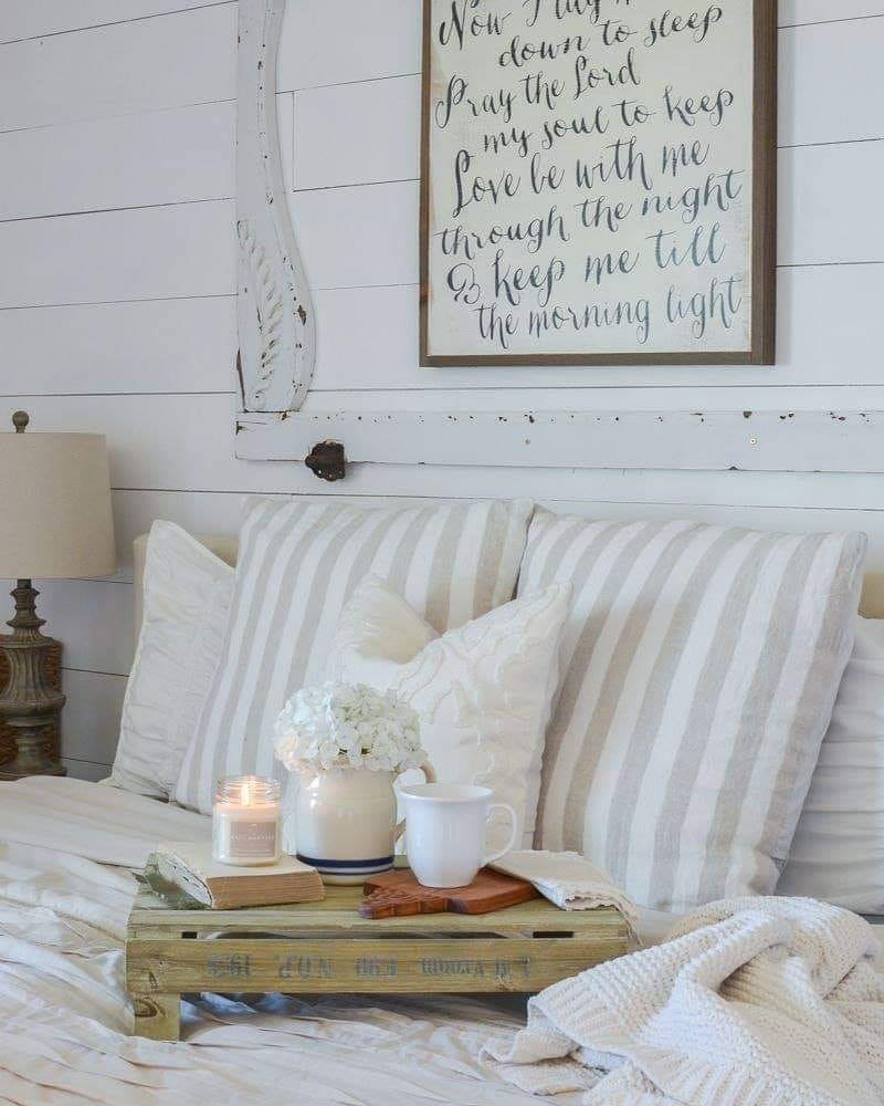 A cozy bedroom scene with striped pillows on a white bed, a tray holding a white vase with flowers, a candle, a cup, and a book sits next to a decorative sign with handwritten text on the wall. Consider incorporating elements from "10 Wooden Crates Decor Hacks to Add Rustic Charm to Your Home" for added warmth.