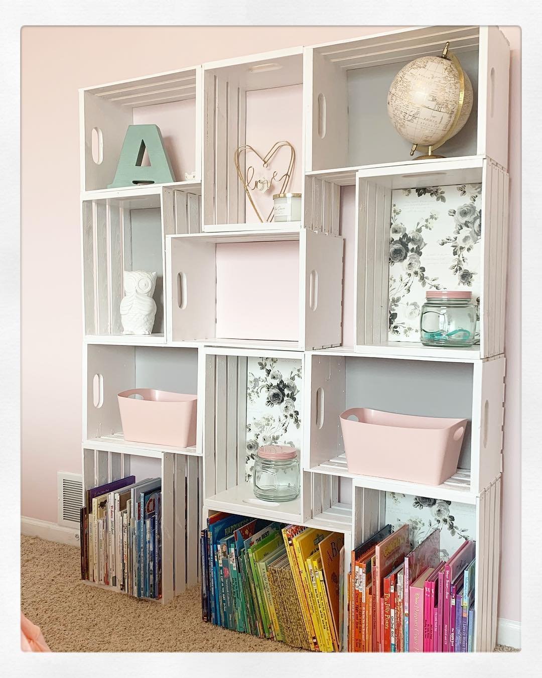 A white wooden storage unit with nine cubby holes contains various items: books, a globe, jars, bins, a heart frame, and decorative letters. Floral wallpaper backs some cubbies. Books line the bottom rows. Discover 10 Wooden Crates Decor Hacks to Add Rustic Charm to Your Home!