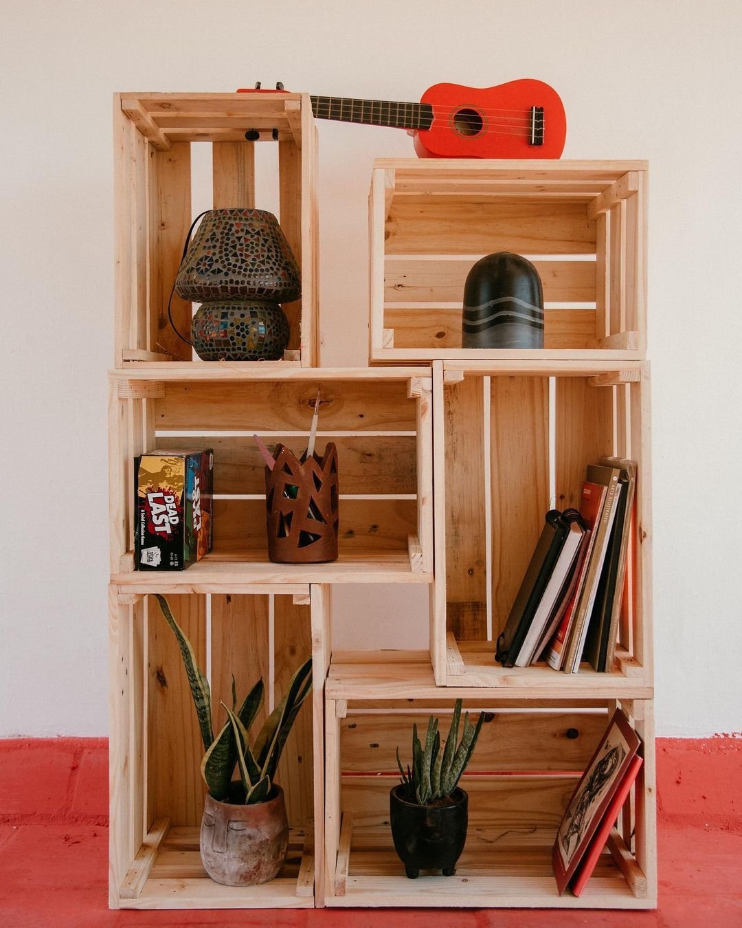 Discover one of the 10 Wooden Crates Decor Hacks to Add Rustic Charm to Your Home: stack wooden crates to create a makeshift shelf holding various items, such as a small lamp, books, potted plants, a puzzle box, and a red toy guitar.