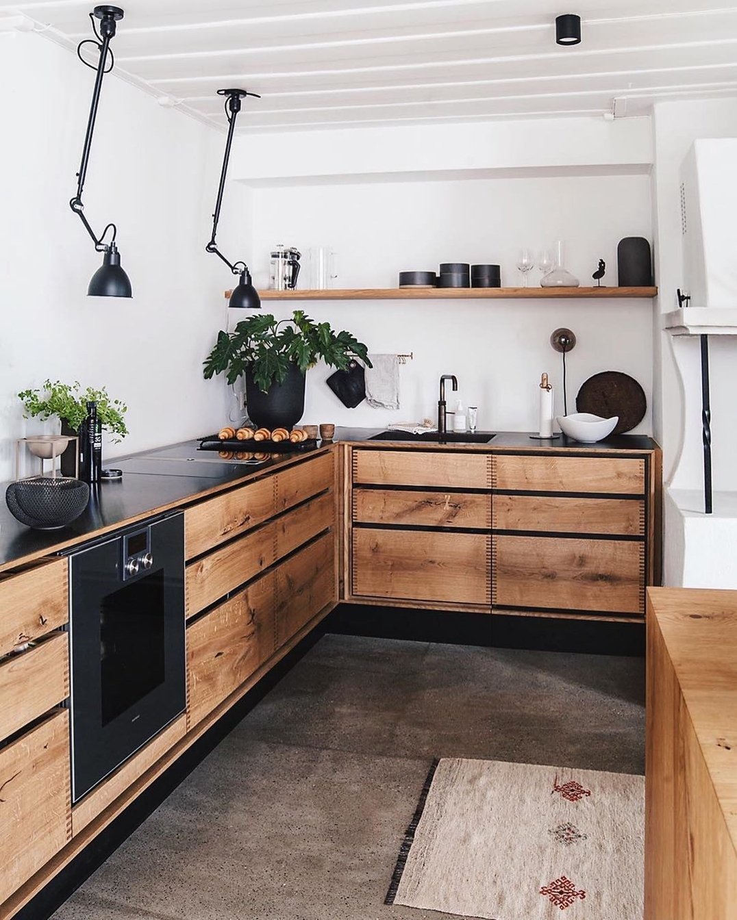 Industrial Design Unveiled: Modern kitchen with wooden cabinets, black countertops, built-in oven, wall-mounted light fixtures, and open shelving adorned with kitchenware and plants. A rug complements the gray floor while white walls and ceiling enhance the space's ultimate decorative charm.