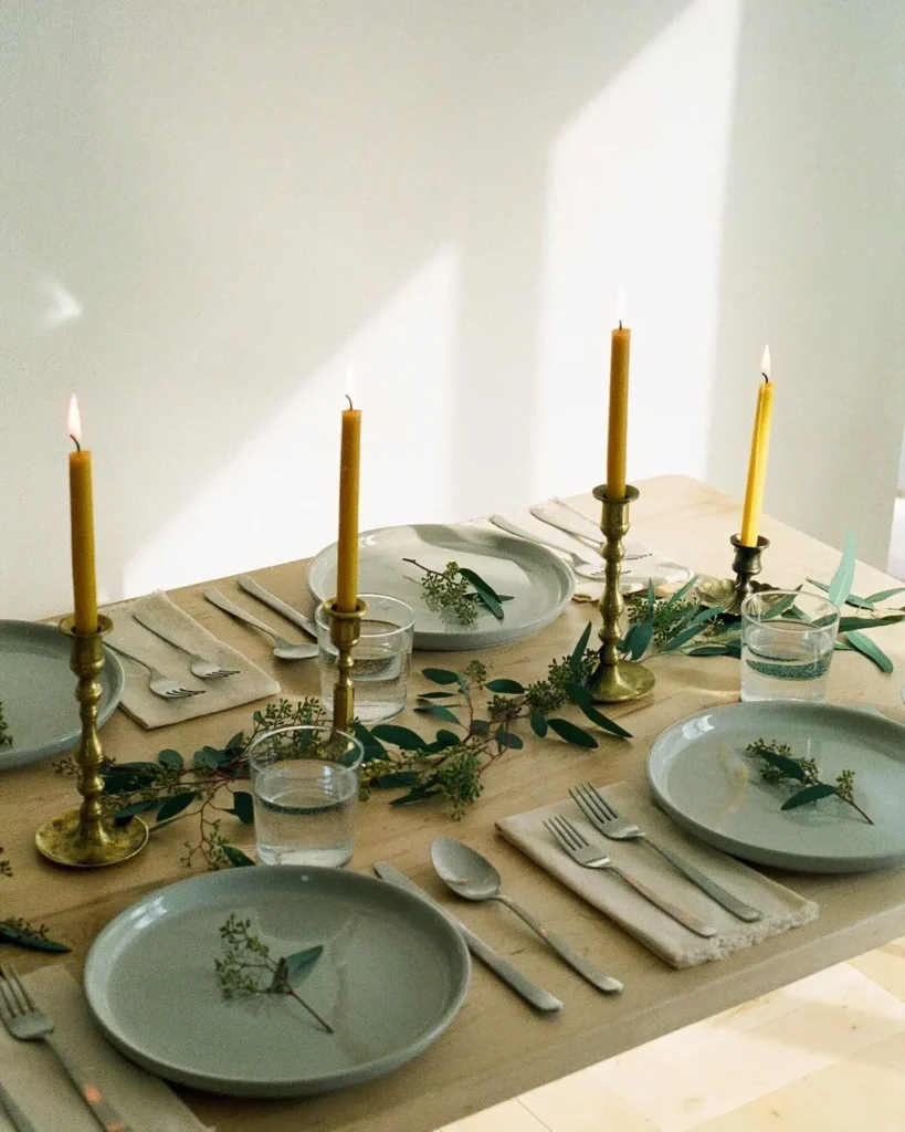 A dining table set for four showcases Minimalist Magic: white plates, silver cutlery, glasses of water, yellow candles in brass holders, and green foliage for a simplified winter table decor.