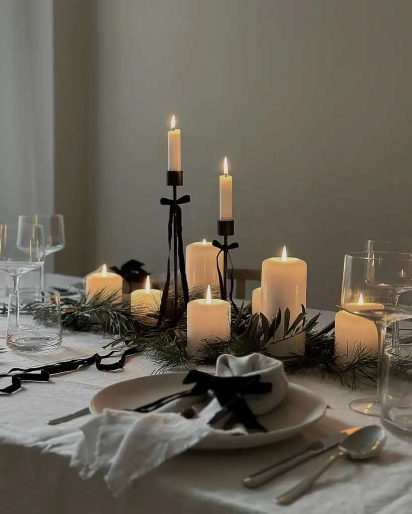 A dining table set with white plates and glasses, decorated with lit candles, black ribbons, and greenery on a white tablecloth brings minimalist magic to your winter table decor.