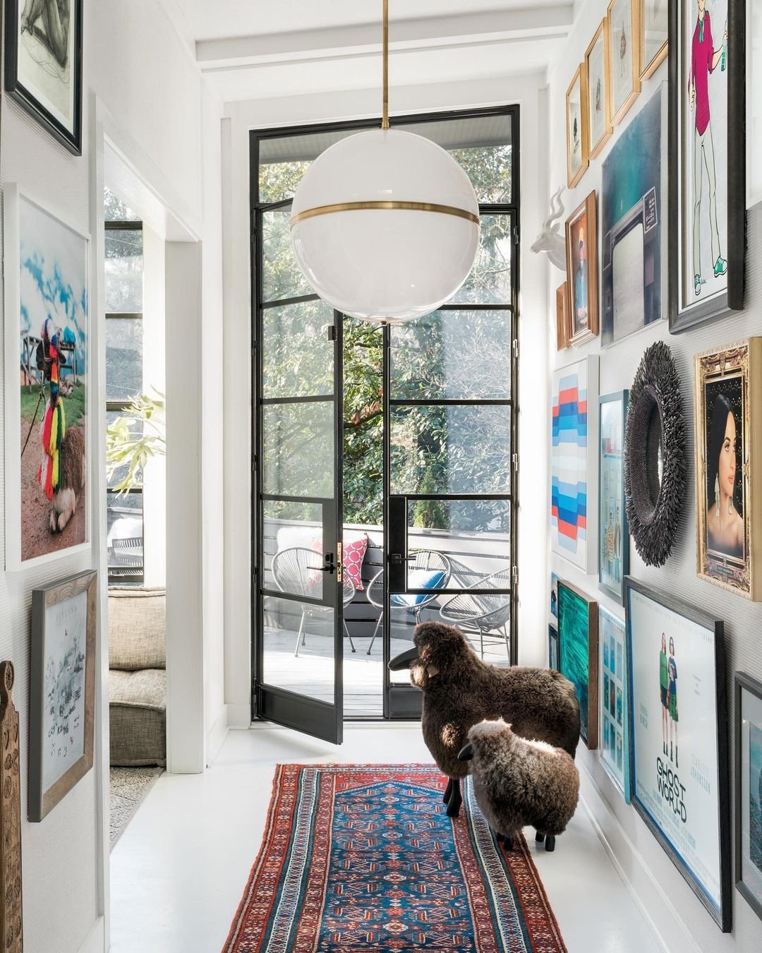 A bright hallway features a steel front door in an industrial style, leading outside. The walls are adorned with various artworks, a patterned rug adds charm, and two plush sheep sculptures complete the chic home design.
