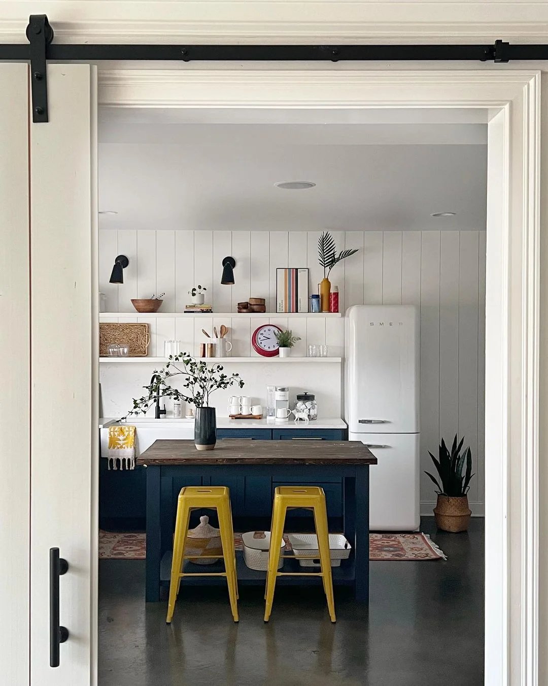 A modern kitchen with white walls, a wooden island with two yellow stools, open shelves with various decor, and a Smeg refrigerator that adds retro style.