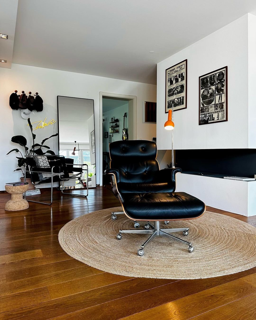 A modern living room unlocking mid century modern magic features a black leather lounge chair on a circular rug, a floor lamp, wall art, a full-length mirror, and a plant. A side table holds a hat collection on the wall.