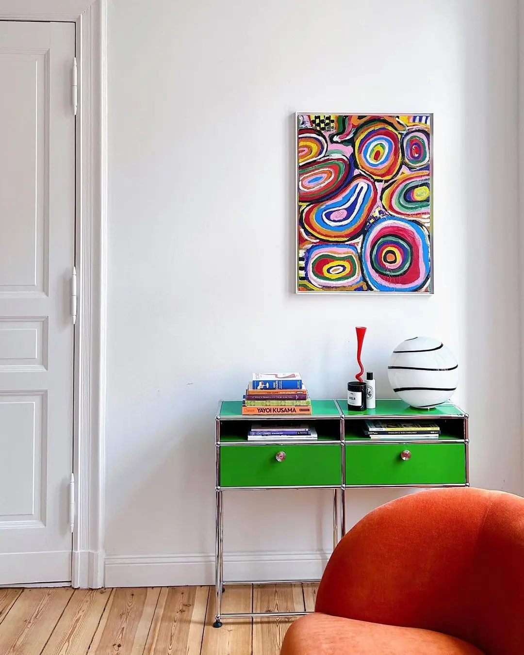 A small green table with books, a white striped lamp, and decor sits against a white wall with abstract colorful art above it—showing How to Add Funky Wall Art to Your Home; a red chair is in the foreground.