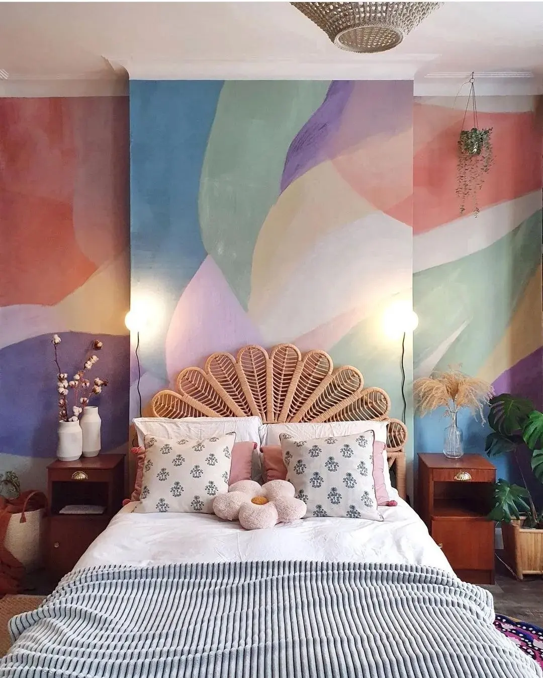A bedroom with a rattan headboard, colorful abstract painted wall, white bedding, funky furniture, decorative pillows, bedside tables, lamps, and various plants—unique pieces to make your home stand out.