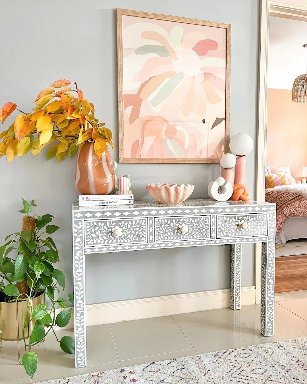 A decorative console table with white floral inlay, fall leaves in a vase, a plant, books, and modern decor stands beneath an abstract art piece on a light gray wall—perfect for fans of Funky Furniture: Unique Pieces to Make Your Home Stand Out.