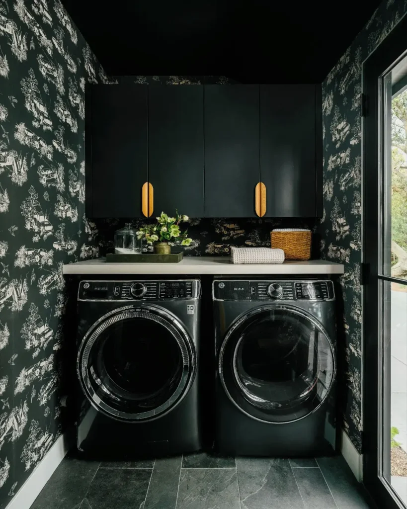 A moody laundry room with black wallpaper, matching black washer and dryer, and black cabinets with gold handles. A countertop adorned with plants adds freshness, while a wicker basket completes the look. Perfect inspiration on how to create a moody laundry room.