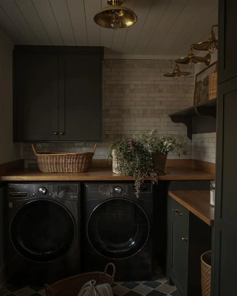 Discover how to create a moody laundry room featuring stacked washing machine and dryer, wooden countertops, a wicker basket, and potted plants. Dark green cabinets seamlessly blend with the white subway tile backsplash and brass fixtures for an inviting yet sophisticated space.