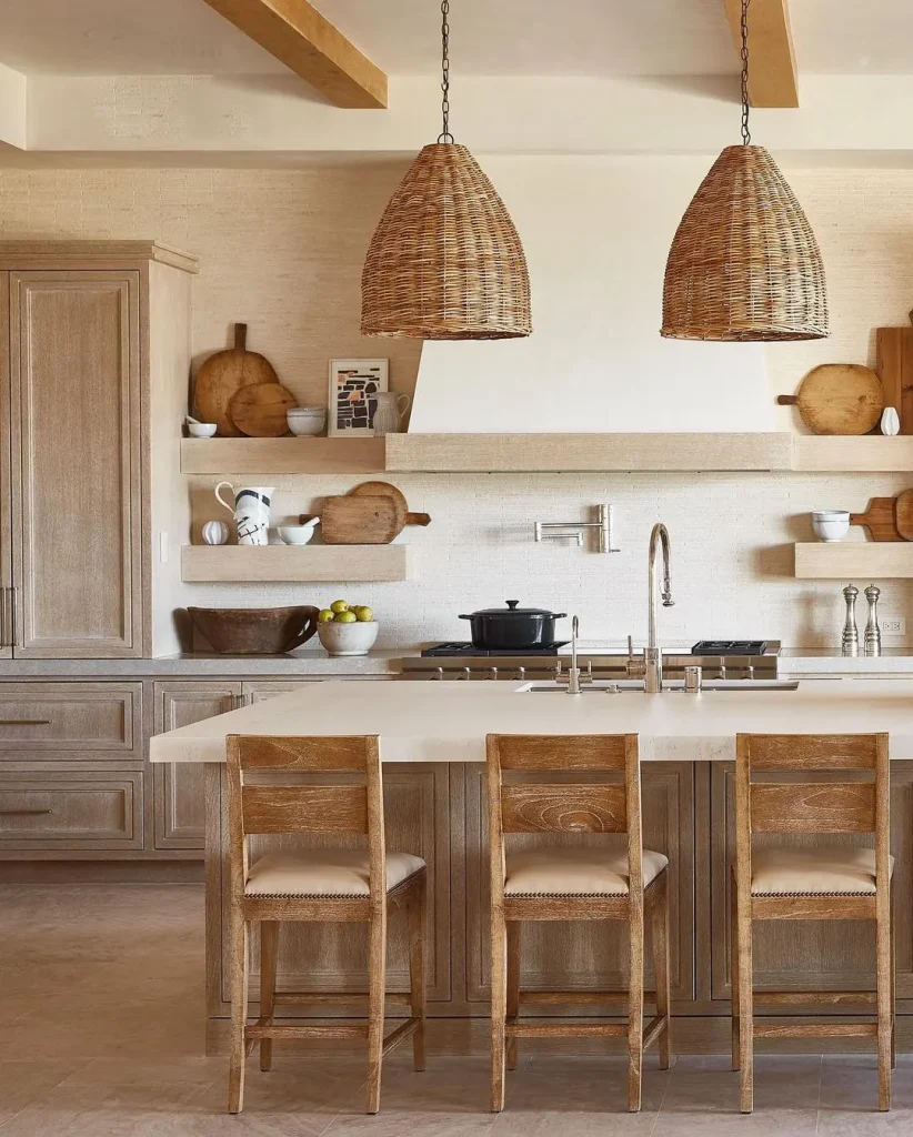 A modern kitchen with wooden cabinets, countertops, and stools. Two woven pendant lights hang above the island, which is equipped with a sink and stovetop. Channeling Beach Decor 101: Everything You Need to Know to Get Started, the decor includes bowls, cutting boards, and jars.