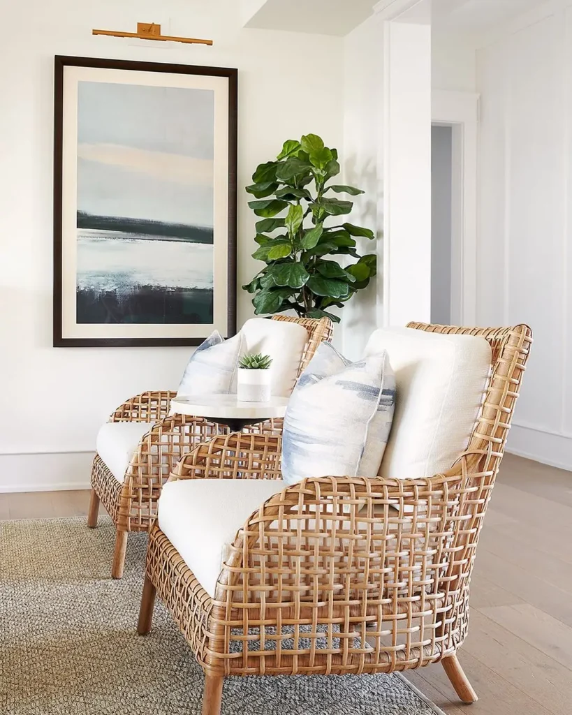 Two wicker armchairs with white cushions and pillows sit by a potted plant and large framed artwork, creating a bright, modern space—perfect inspiration for How to Design a Beach-Styled Living Room on a Budget.