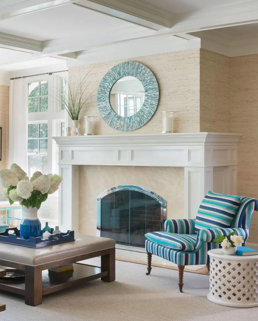 A living room with a striped armchair, a round mirror above the fireplace, and a coffee table with white flowers in a blue vase subtly showcases beach decor: how to select colors and materials for your home. The room has neutral walls, light carpet, and large windows.