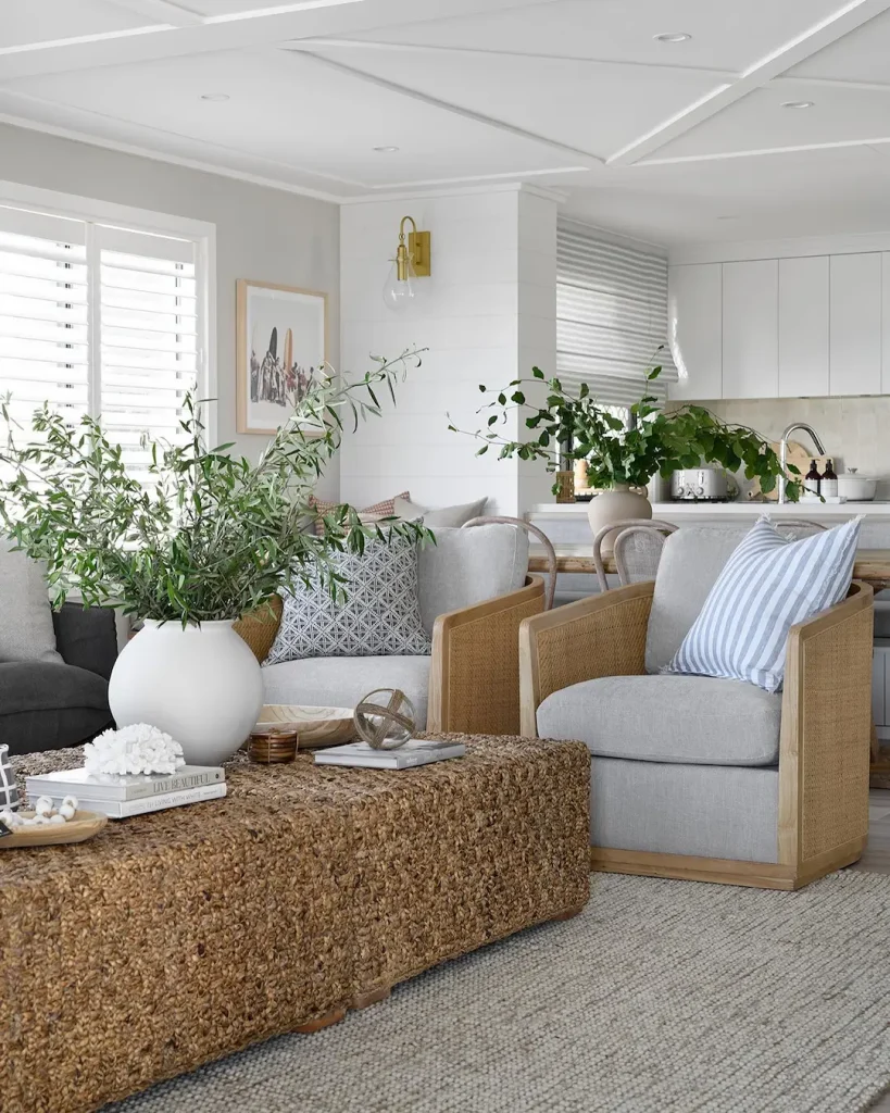 Bright living room with neutral-toned furniture, a woven coffee table, potted plants, and an open view into a white kitchen—perfect inspiration for Beach Decor 101: Everything You Need to Know to Get Started.