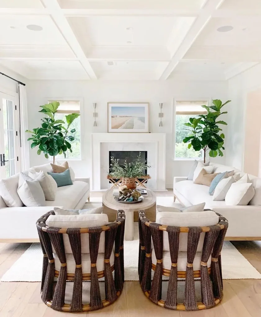 A bright living room with two white sofas, two woven armchairs, a coffee table, a white fireplace, green plants, and a landscape painting above the mantel. This setup is perfect for those looking to learn how to design a beach-styled living room on a budget.