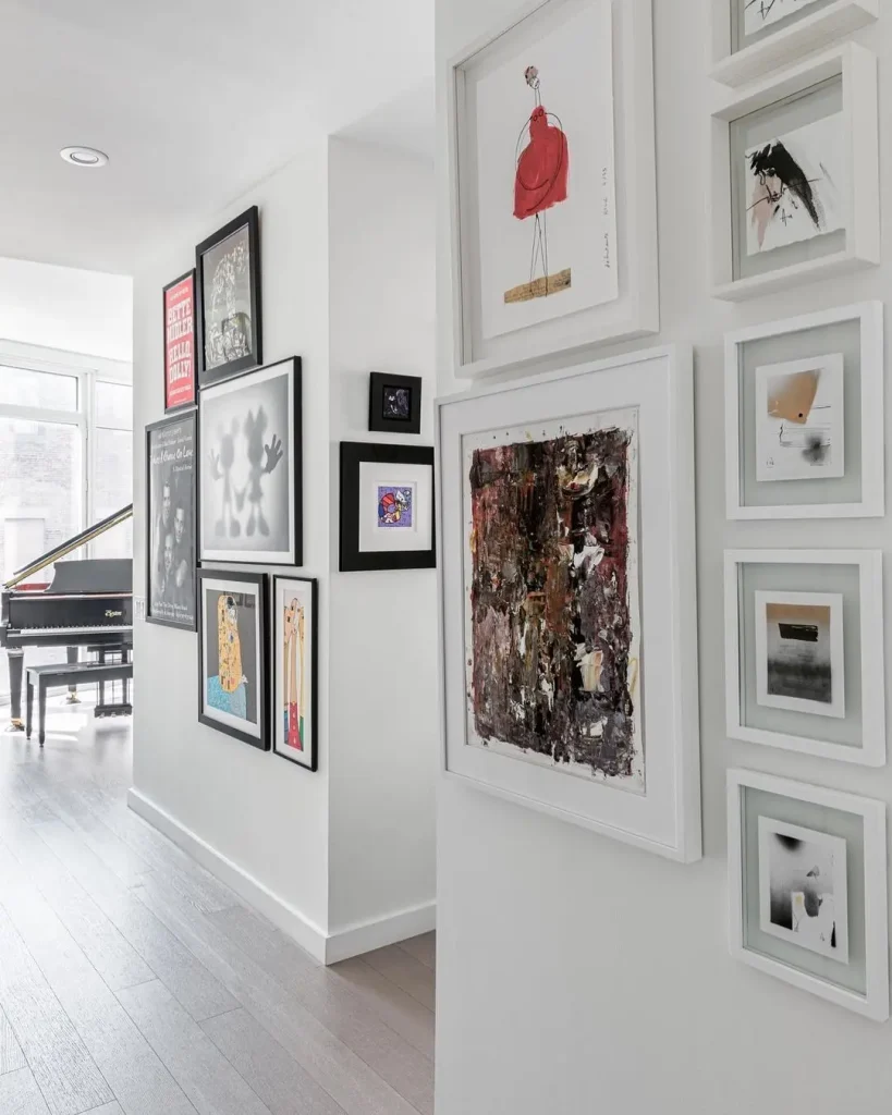 A hallway showcasing a stunning gallery wall with framed art leads to a bright room with a grand piano by large windows.