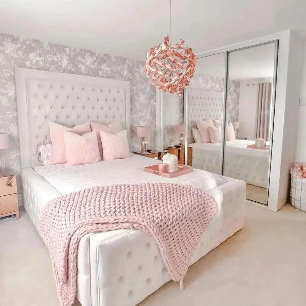 A bedroom featuring a large bed with a tufted headboard, pink pillows, and a pink knit throw. A mirrored closet, wall-mounted lamps, and a decorative light fixture are also present. Follow these tips on how to create a whimsical pink master bedroom retreat that exudes charm and elegance.