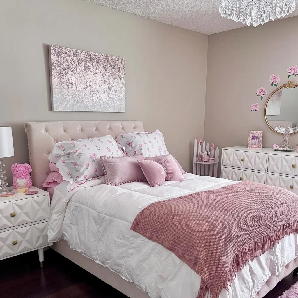 A neatly made bed with pink and white bedding, floral pillows, and a pink blanket sits in a pastel-themed bedroom with a chandelier, dresser, and wall art—showcasing how to create a whimsical pink master bedroom retreat.