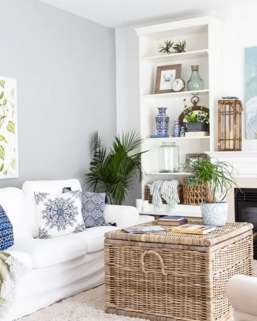 A bright living room with a white sofa, blue and white patterned cushions, wicker coffee table, green plants, and decor-filled shelves shows how to design a beach-styled living room on a budget.