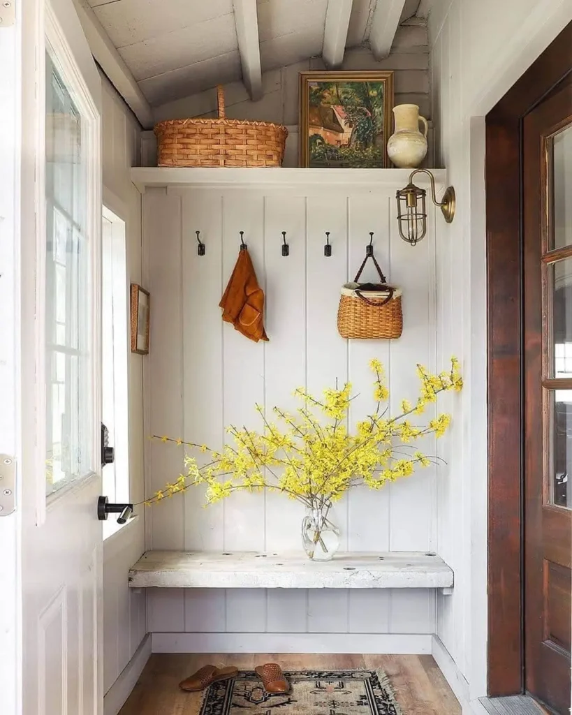 A small entryway, your ultimate functional and stylish mudroom guide, features a wooden bench, yellow flowers in a glass vase, a cozy rug, and hanging hooks. A shelf above displays a basket, picture, and vase. A brown door stands to the right.