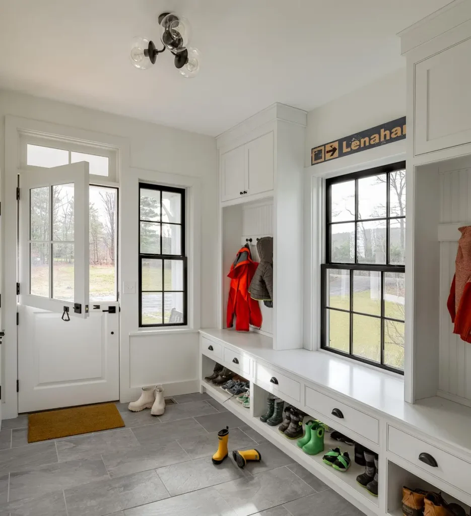 Discover your ultimate functional and stylish mudroom guide with white cabinetry, a convenient bench, and hooks for hanging coats. Boots find their place below, while natural light streams through a window and door. A small mat adds charm on the tiled floor.