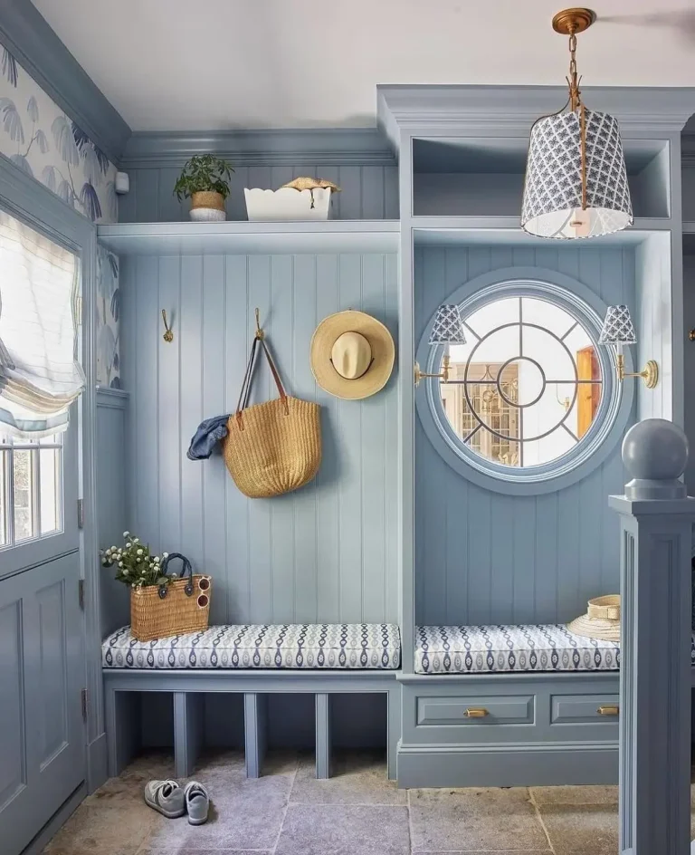 A tidy blue mudroom with built-in benches, hooks holding hats and a straw bag, a round window, patterned cushions, and decorative items on upper shelves—discover ideas in Your Ultimate Functional and Stylish Mudroom Guide.