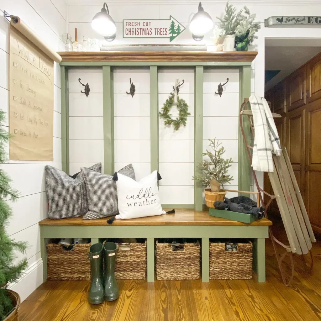A mudroom bench with three cubbies containing baskets, coat hooks above, holiday decorations, pillows, rain boots, and a sled leaning against the wall—discover inspiration in Your Ultimate Functional and Stylish Mudroom Guide.
