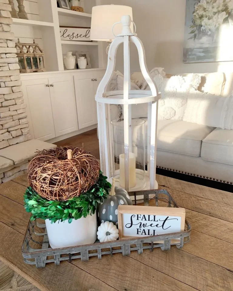 A decorative tray on a wooden table showcases how to style your fall farmhouse coffee table, featuring a faux pumpkin, greenery, lantern, "Fall Sweet Fall" sign, and other autumn accents in a cozy living room.