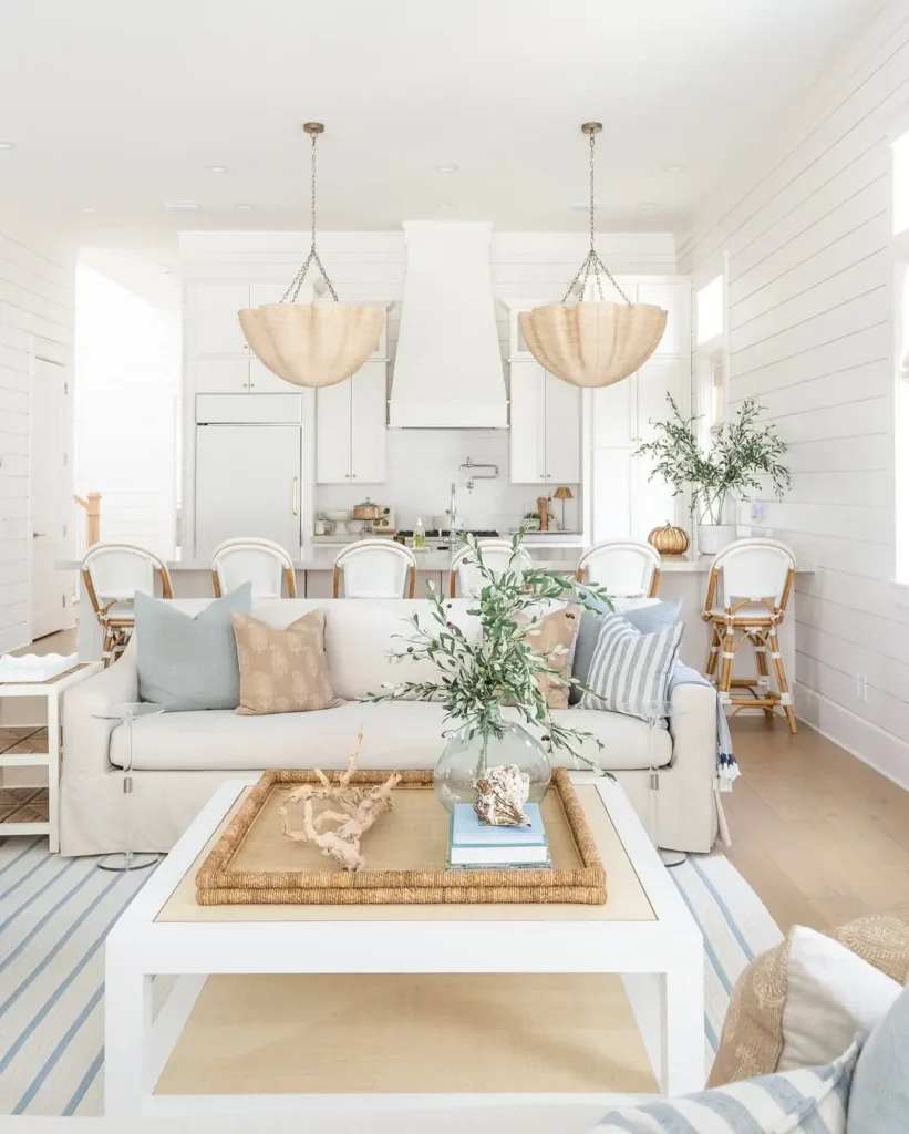 Bright, airy coastal living room and kitchen with neutral decor, a white sofa, striped rug, wooden accents, and pendant lights over a dining table with white chairs.
