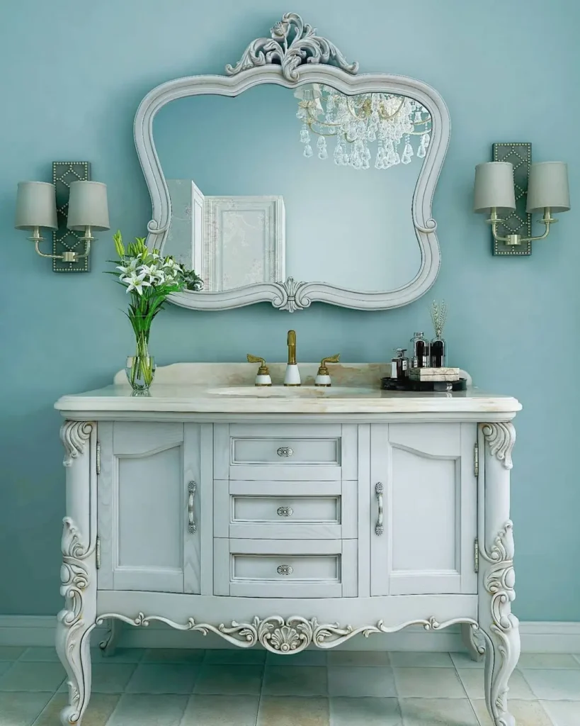 A vintage-style bathroom vanity with ornate details, a marble countertop, a french country mirror, floral arrangement, and wall sconces on a light blue wall.