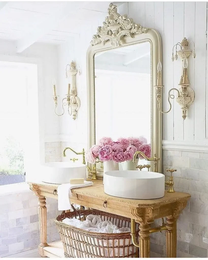 A bathroom with a large french country mirror, double sinks on a wooden vanity, gold fixtures, wall sconces, a basket below, and a vase of pink flowers on the counter.