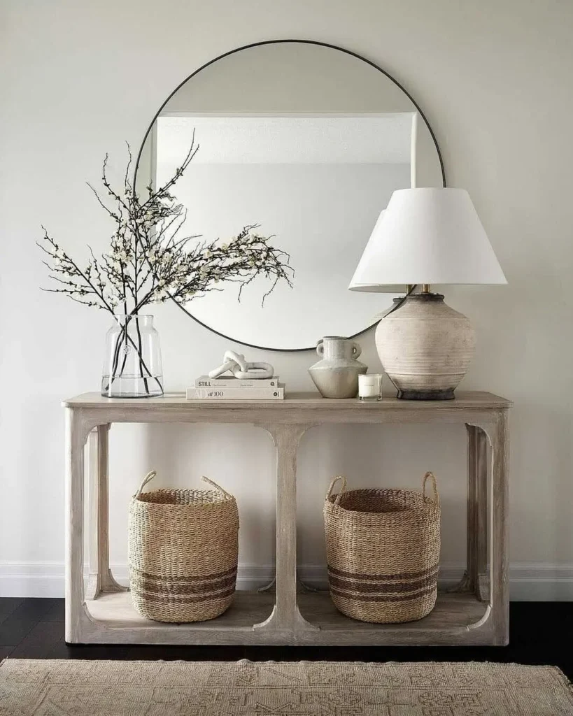 A round mirror hangs above a rustic console table in light wood, styled with a lamp, a vase of branches, books, and two woven baskets on the lower shelf.