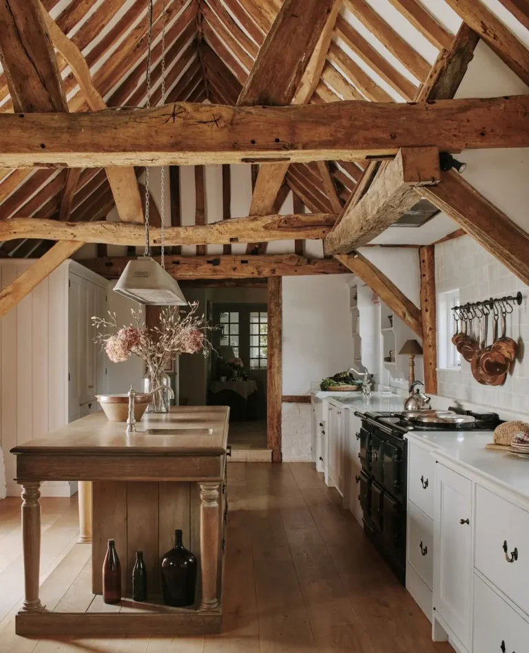 A rustic kitchen with exposed wooden beams, a wooden island with vases, white cabinets, a black stove, and hanging utensils on a white tile wall, perfect for inspiring your rustic kitchen ideas.