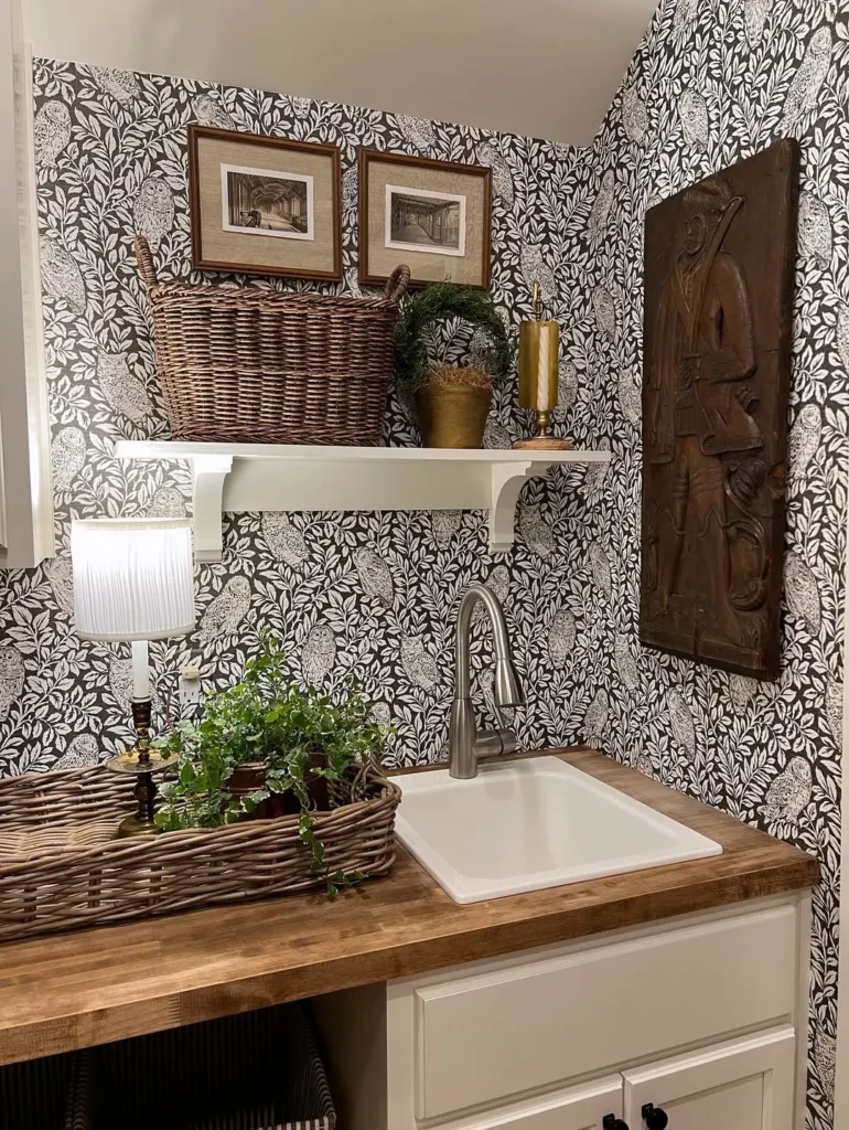A small sink area with a wood countertop, black-and-white floral wallpaper to add character to a bland all-white laundry room, decorative baskets, framed art, potted plants, and a wall-mounted metal relief artwork.