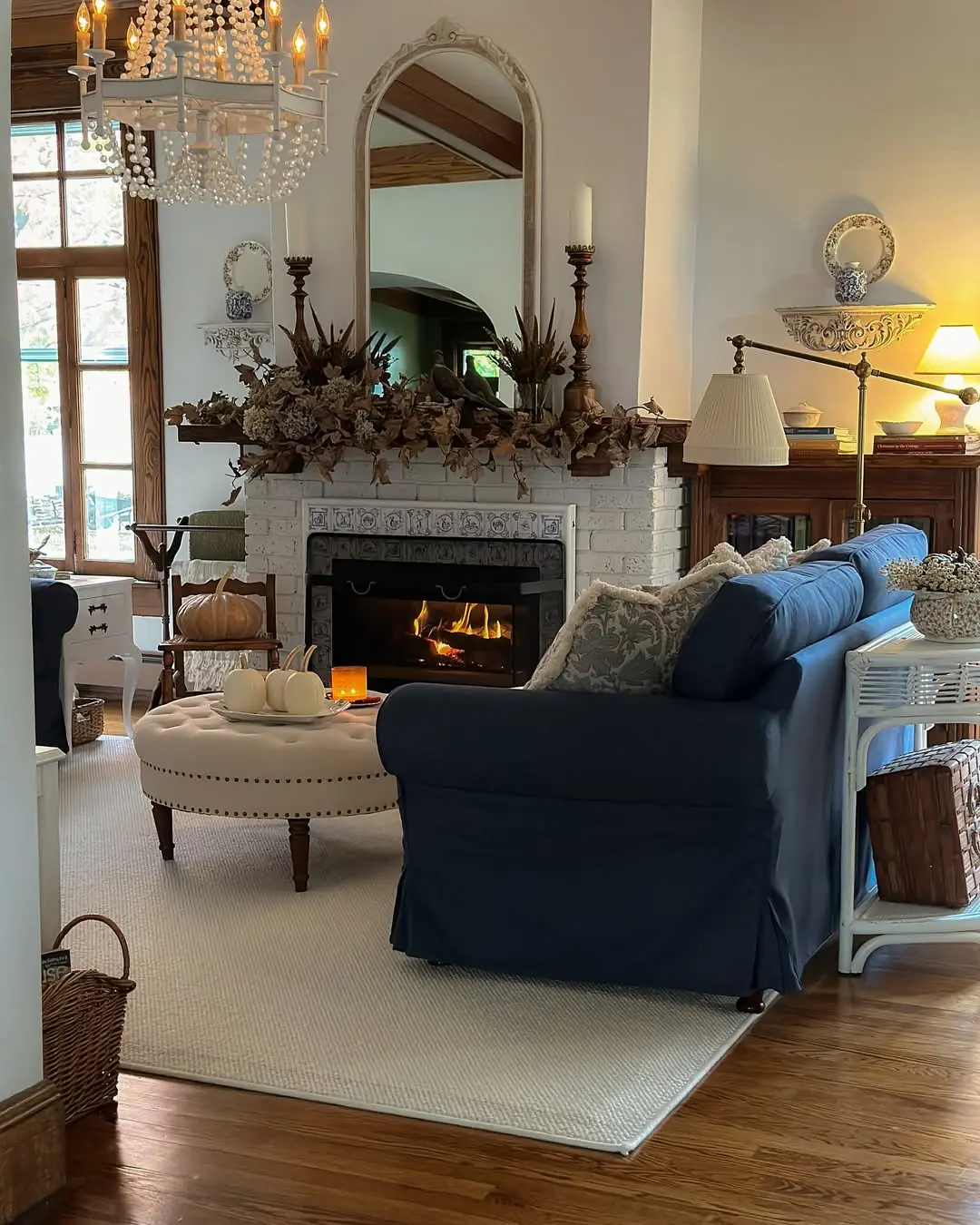 A cozy fall living room with a lit fireplace, white pumpkins on a tufted ottoman, warm candlelight, and autumn floral arrangements creating a welcoming seasonal atmosphere.