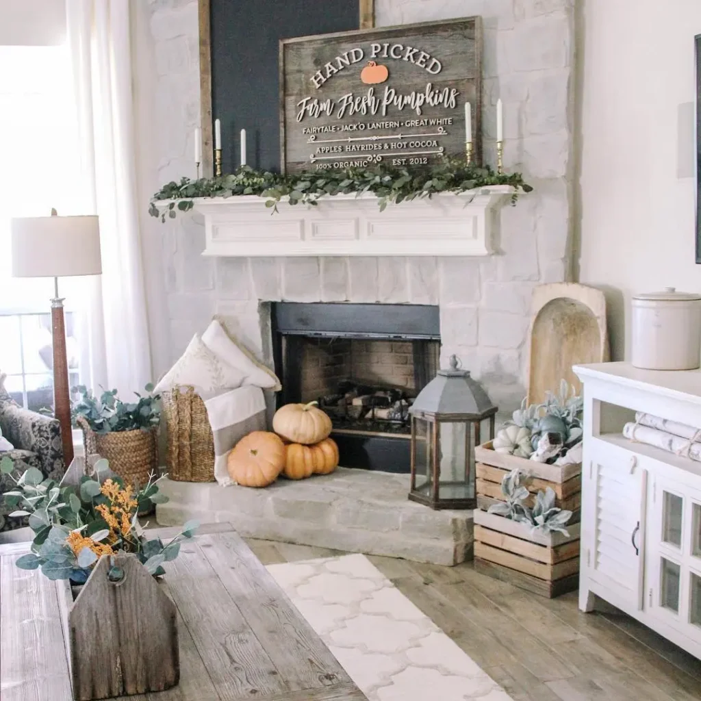A stone fireplace decorated with pumpkins, greenery, candles, a lantern, and a rustic sign creates a bright, cozy living room perfect for rustic fall charm.