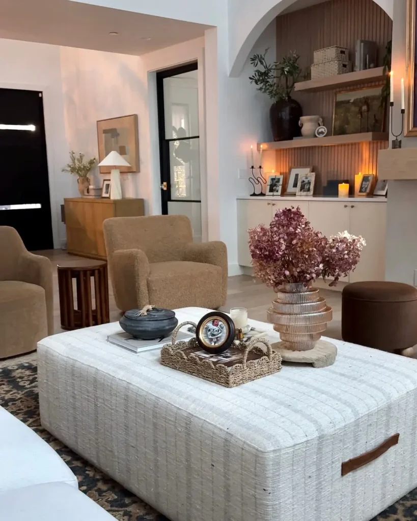 Organic modern living room styled for fall featuring warm neutral tones, textured armchairs, a woven tray on a linen ottoman, dried flowers in a glass vase, and soft candlelight for a cozy, natural feel.