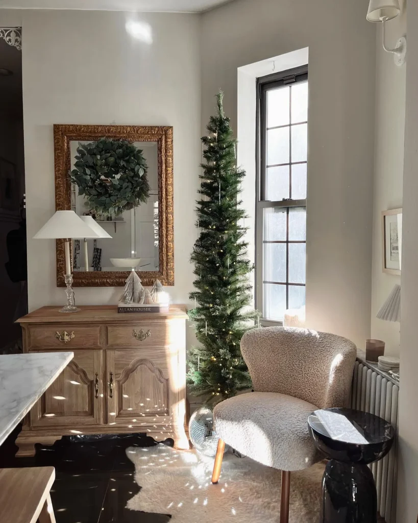 A small pencil Christmas tree stands next to a window in a bright corner with a wooden sideboard, framed mirror, white lamp, and a textured armchair.