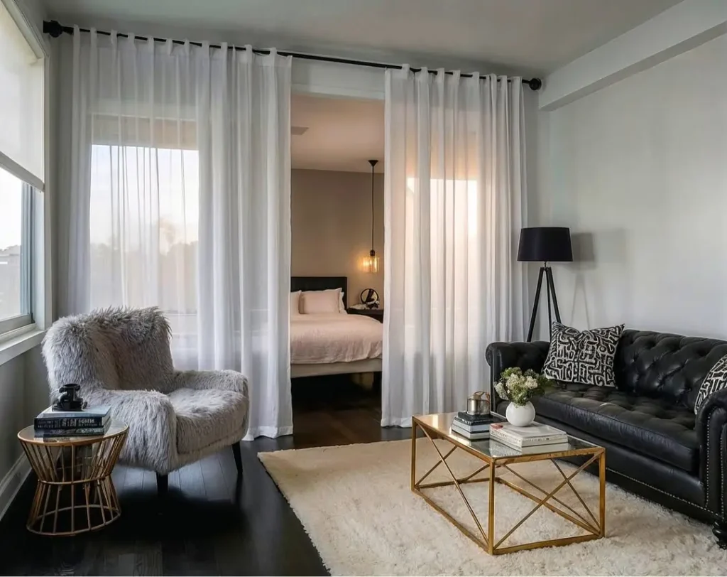 Modern living room with a black leather sofa, fur accent chair, and gold coffee table defines studio apartment zones, while sheer curtains open to a bedroom area with a bed and pendant light.