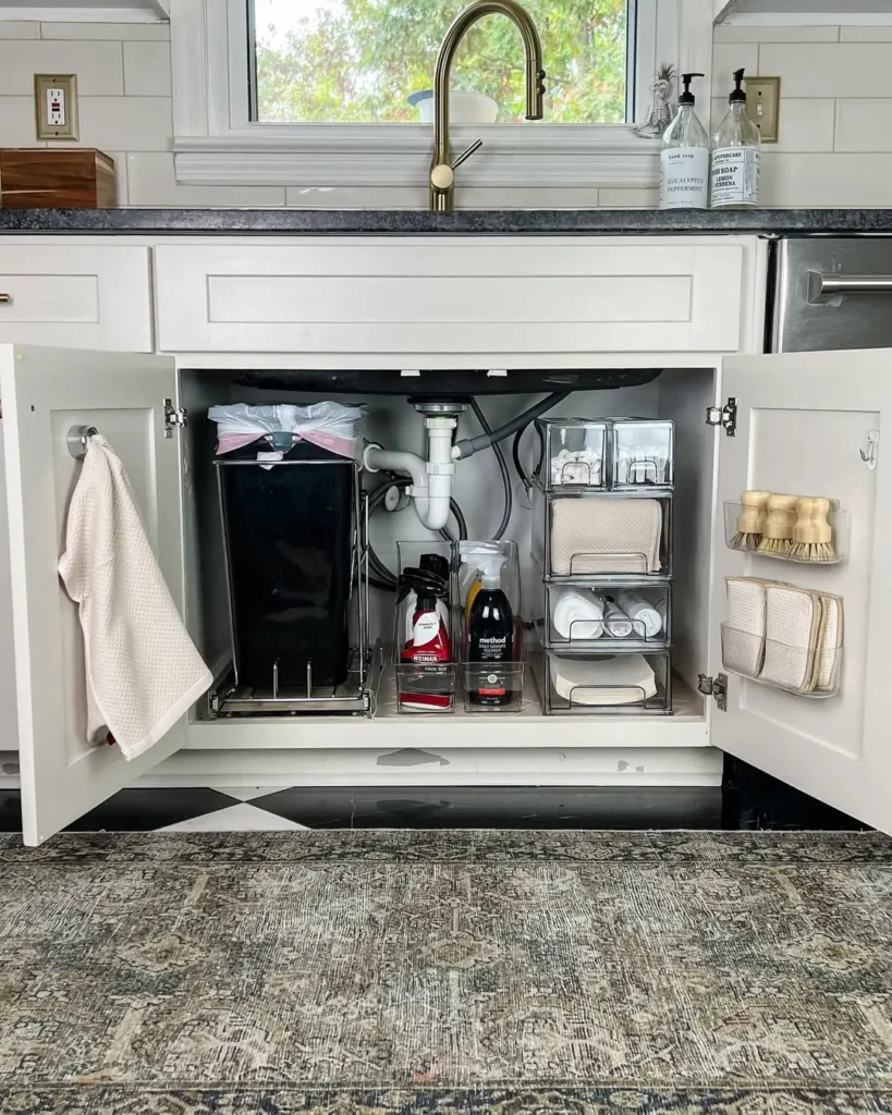Open kitchen cabinet under a sink, organized with cleaning supplies, towels, storage bins, and brushes on shelves and racks.
