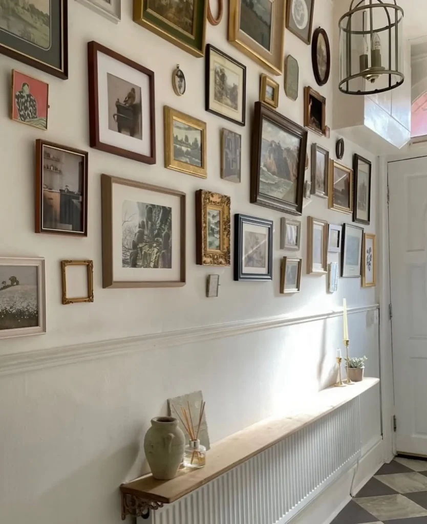 A hallway with white walls features a striking gallery wall of framed art and photos. A narrow shelf holds a vase, candles, and diffuser near a white door beneath a hanging lantern.